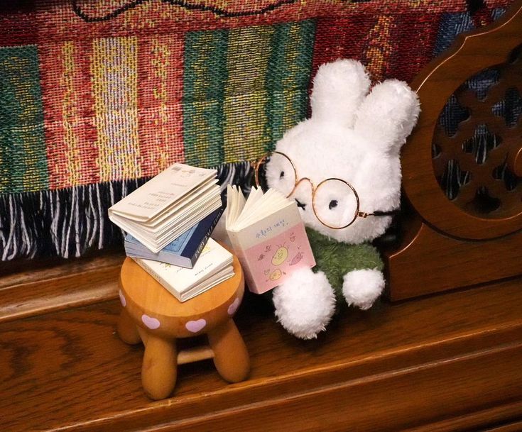 A small miffy plushie wearing round wire rimmed galsses and a green sweater. She is sitting at a small round wooden table reading a book with a pile of three more books infront of her.